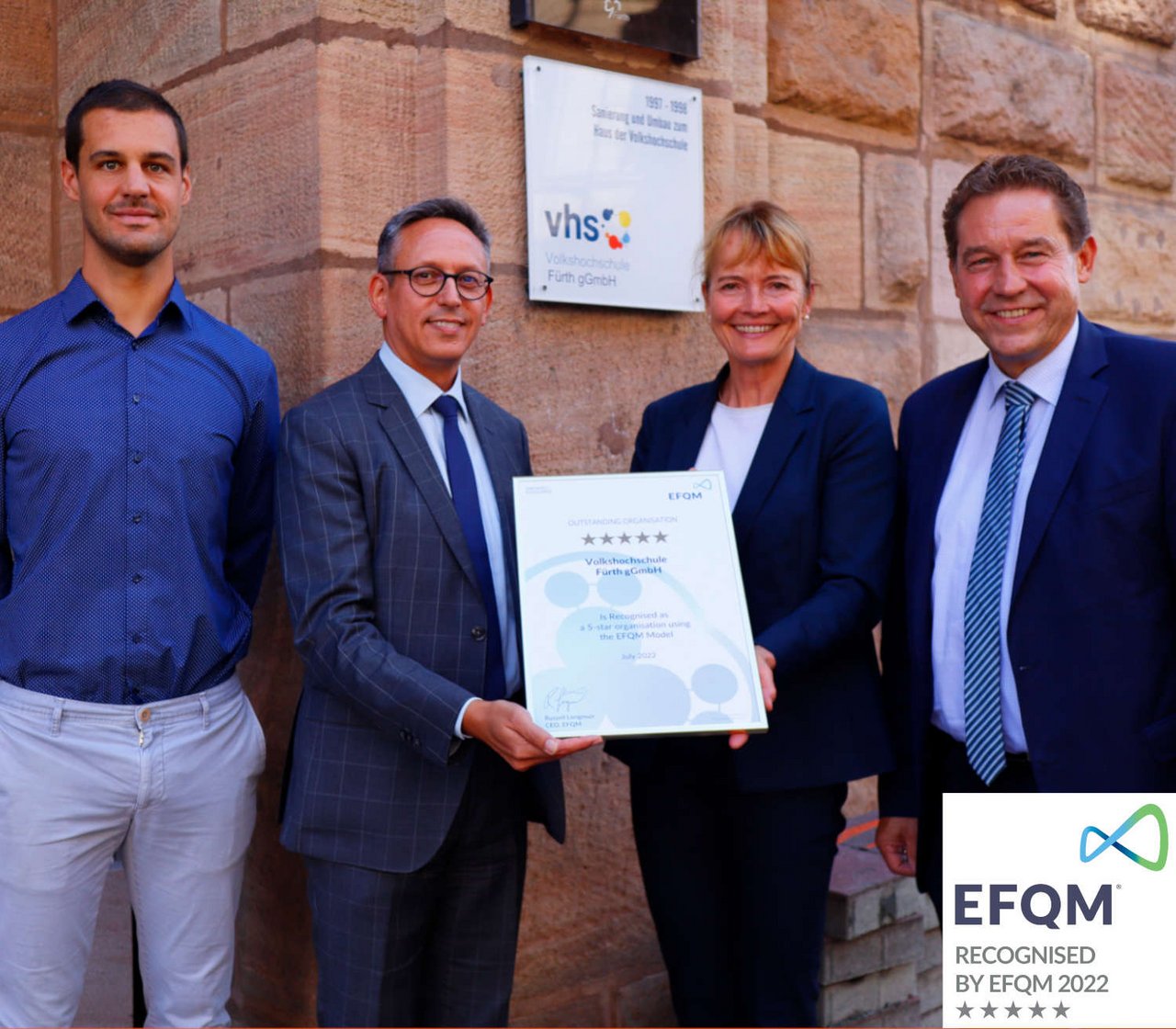 Vier Personen stehen vor der vhs. Der Leiter hält ein Zertifikat hoch.